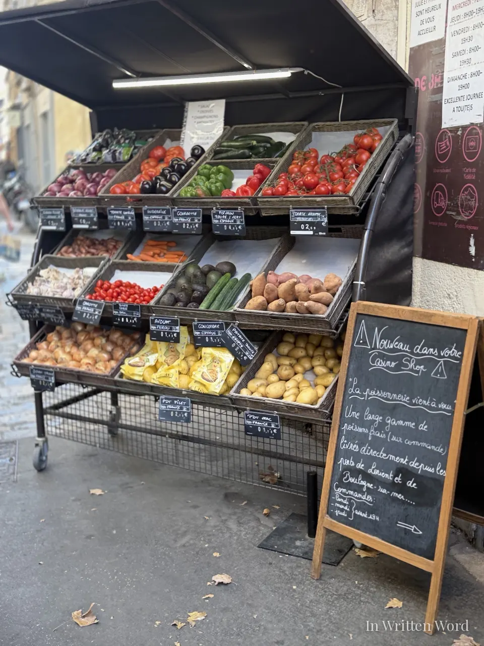 Wicker baskets of tomatoes, peppers, avocados and potatoes on a sidewalk produce stand beside a handwritten chalkboard in Aix-en-Provence.