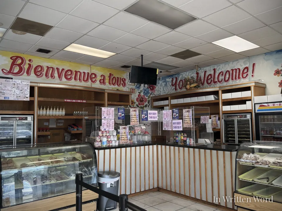 Interior of Piman Bouk Bakery in Miami showing the service counter, glass display cases, and painted signs reading “Bienvenue à tous” and “You Welcome!” above the counter.