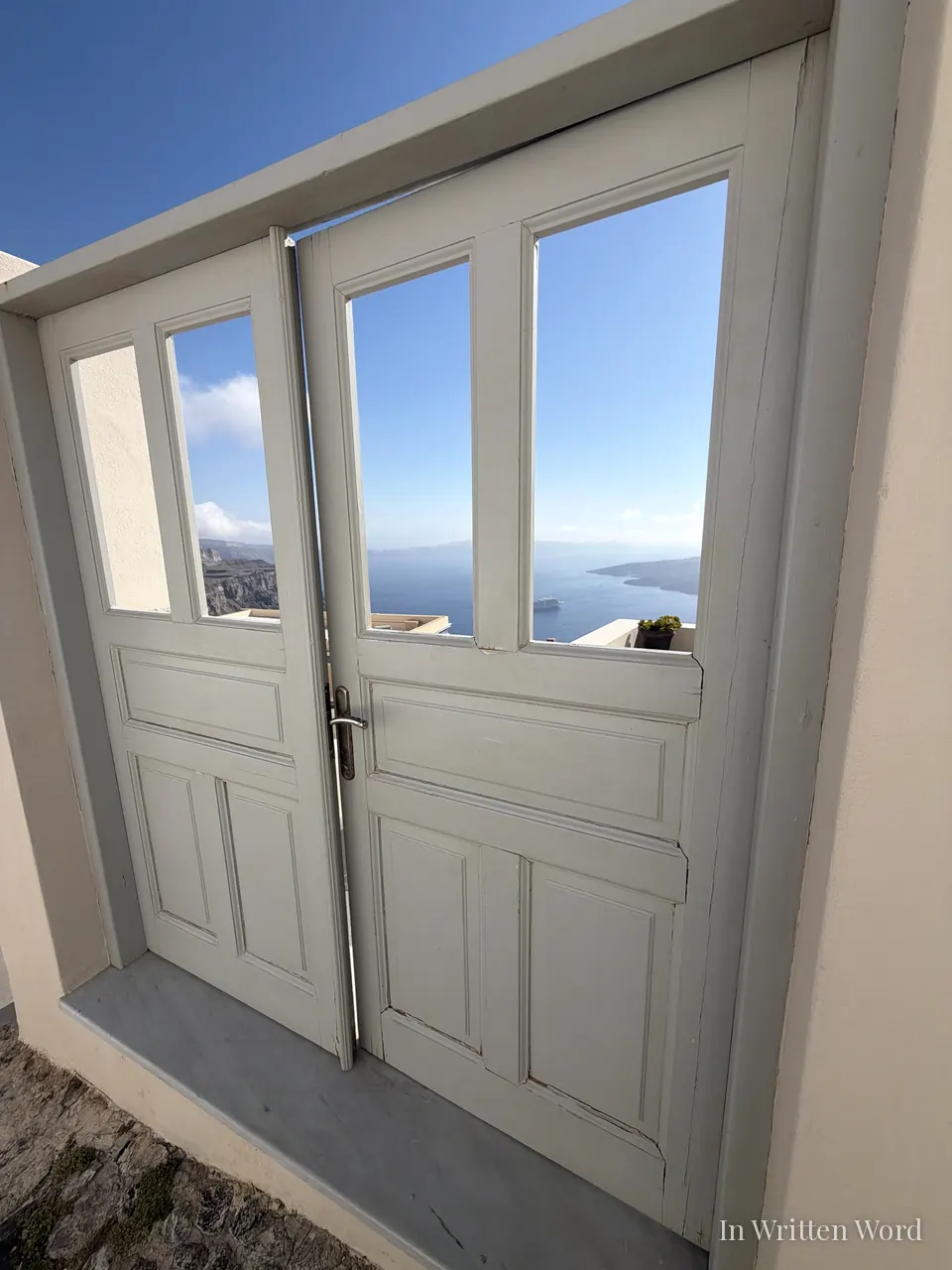 Two pale wooden doors with no glass panes opening onto a wide view of the Santorini caldera, the deep blue Aegean, and the distant cliffs of the opposite shore.