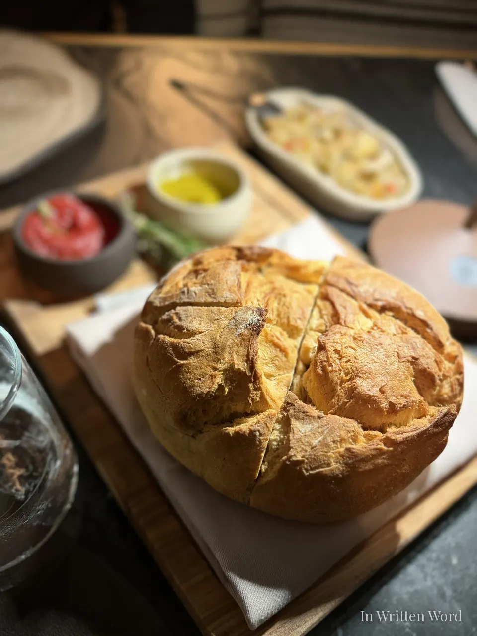 A rustic loaf of crusty Greek bread on a wooden board, accompanied by small bowls of olive oil, red pepper spread, and seasonal sides in soft natural light.