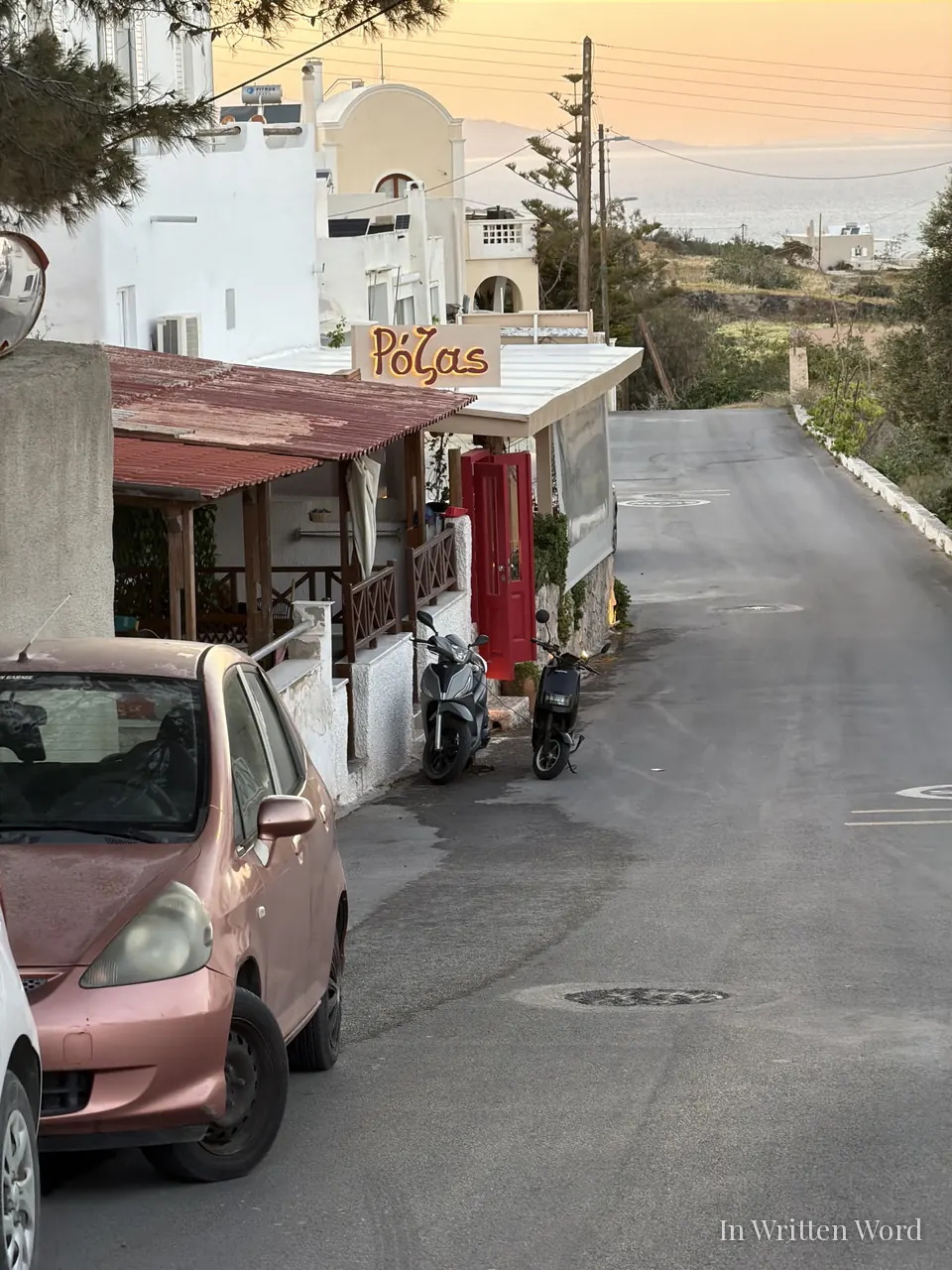 A small Santorini taverna at golden hour, with a red door, a vine-covered awning, two scooters outside, and the Aegean Sea visible in the distance down a quiet sloping street