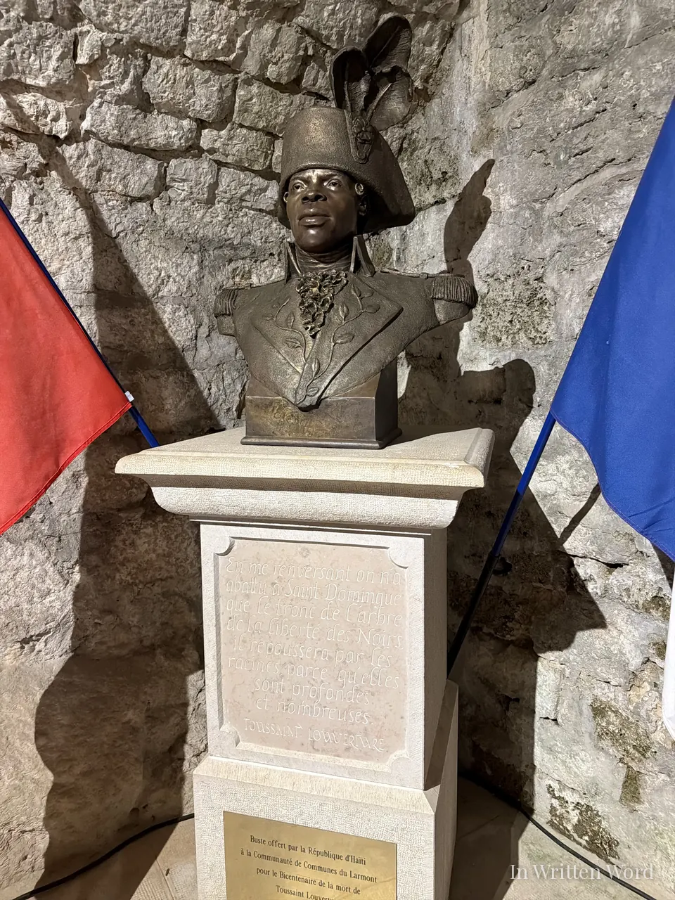 A bronze bust of Toussaint L'Ouverture on a stone plinth at Fort de Joux, flanked by the Haitian and French flags, with an engraved French quote below.